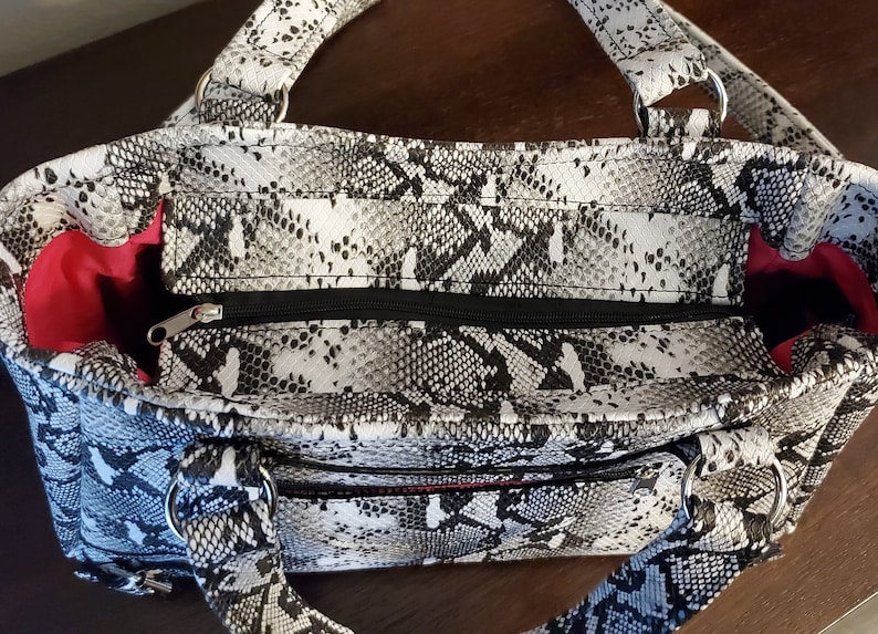 May include: A black and white snakeskin print handbag with a zipper closure and two silver rings on the handles. The bag has a red lining.