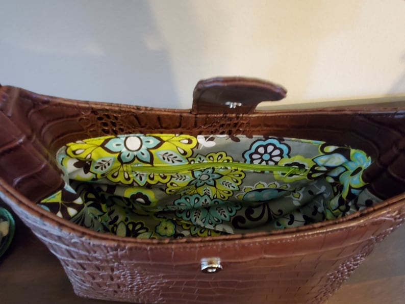 May include: A brown faux crocodile leather handbag with a floral patterned lining. The bag has a top flap closure with a silver buckle.