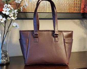 May include: A brown leather tote bag with two top handles. The bag has a zipper closure and a pocket on the side.