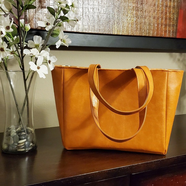 May include: A brown leather tote bag with two straps. The bag is sitting on a wooden table.