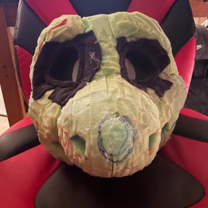 May include: A green and black foam head for a furry costume. The head has two eye holes and a nose hole. The foam is marked with lines and circles.