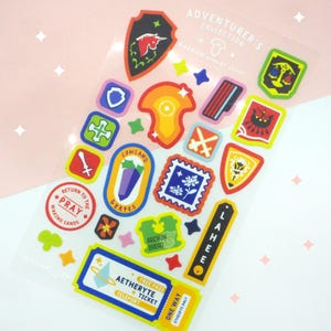 May include: A sheet of colorful stickers featuring various designs, including shields, stamps, and a ticket. The stickers have a fantasy theme, with text like "Adventurer's Collection" and "Aetheryte Ticket". The color palette includes red, blue, yellow, and green.