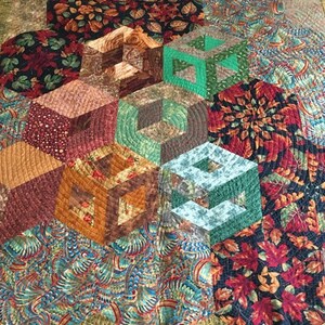 May include: A quilt featuring a geometric pattern of cubes in shades of brown, green, and blue. The quilt is made up of various fabric patterns, including floral and paisley designs.