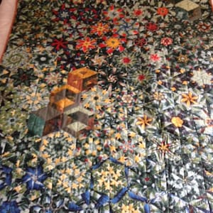 May include: A colourful quilt with a geometric pattern of stars and flowers. The quilt is made of various fabrics in shades of green, blue, yellow, red, and brown.