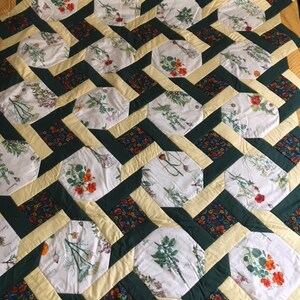May include: A quilt with a geometric pattern of green, yellow and white fabric. The fabric features a floral print with a variety of flowers and plants.