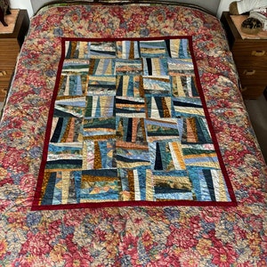 May include: A patchwork quilt with a red border and a pattern of colourful squares and rectangles. The quilt is made of different fabrics and textures, creating a unique and interesting design.