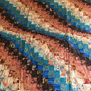 May include: A patchwork quilt in shades of blue, brown, and cream. The quilt is made from various fabrics with floral and geometric patterns. The stitching creates a diagonal design across the surface. The quilt is handmade.