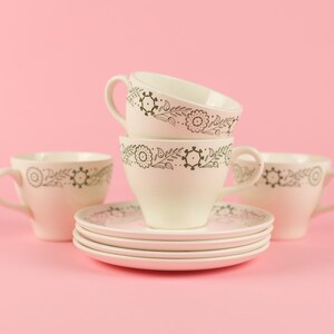May include: A stack of four white teacups with black floral designs on a pink background. The cups are stacked on top of each other, with the top cup being the largest and the bottom cup being the smallest.