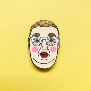 Könnte beinhalten: Emaille-Anstecknadel mit einem stilisierten Gesicht mit heller Haut, blonden Haaren und einer Brille mit weißem Rahmen. Das Gesicht hat blaue Augen, rosa Wangen und einen roten, offenen Mund. Die Anstecknadel befindet sich auf einem hellgelben Hintergrund.