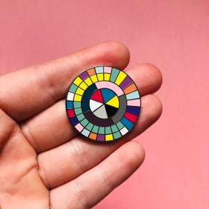 May include: A colorful enamel pin with a geometric design. The pin features a circular pattern of various colors, including red, yellow, blue, and purple. The pin is held in a hand against a pink background. The pin is approximately 1 inch in diameter.