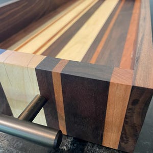 May include: A rectangular wooden tray with a variety of wood tones, including light, medium, and dark browns. The tray has a metal handle and is constructed with vertical and horizontal wood grain patterns. The tray is designed for serving or display.