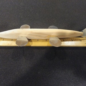 May include: A wooden fish lure with a light brown finish and silver fins. The lure is approximately 12 inches long and is resting on a wooden ruler.