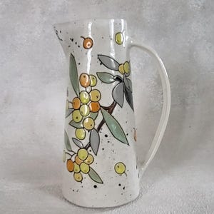 Op de afbeelding: Witte keramische kan met een handvat, versierd met een botanisch ontwerp van gele en oranje bessen, groene bladeren en grijze accenten. De kan heeft een gespikkelde afwerking.