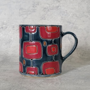 Peut inclure: Une tasse en céramique avec un fond bleu foncé et un motif répétitif de carrés et de rectangles rouges et oranges. La tasse a une anse arrondie et une base beige clair. Le design rappelle l'art moderne du milieu du siècle.