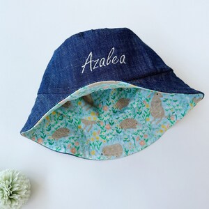 May include: A blue denim bucket hat with a floral print lining. The hat is embroidered with the name "Azalea".