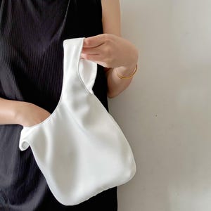 May include: A solid white satin-finish handbag with a single integrated handle. The bag features a soft, unstructured silhouette with a smooth, reflective surface and a simple, minimalist design that hangs from a single loop closure.