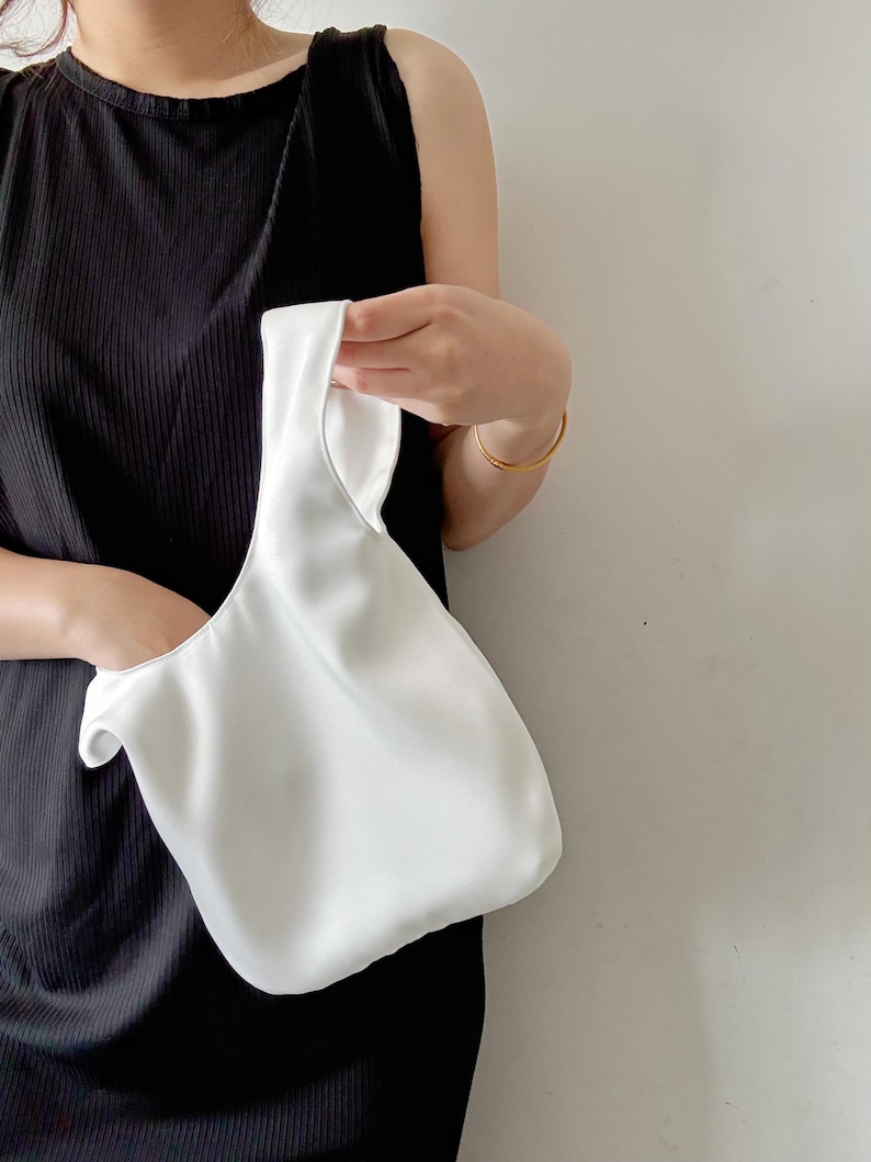 May include: A solid white satin-finish handbag with a single integrated handle. The bag features a soft, unstructured silhouette with a smooth, reflective surface and a simple, minimalist design that hangs from a single loop closure.