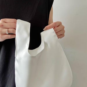 May include: A white tote bag with a rounded shape and two handles. The bag is held up by a person. The bag is made of a smooth, solid white material.