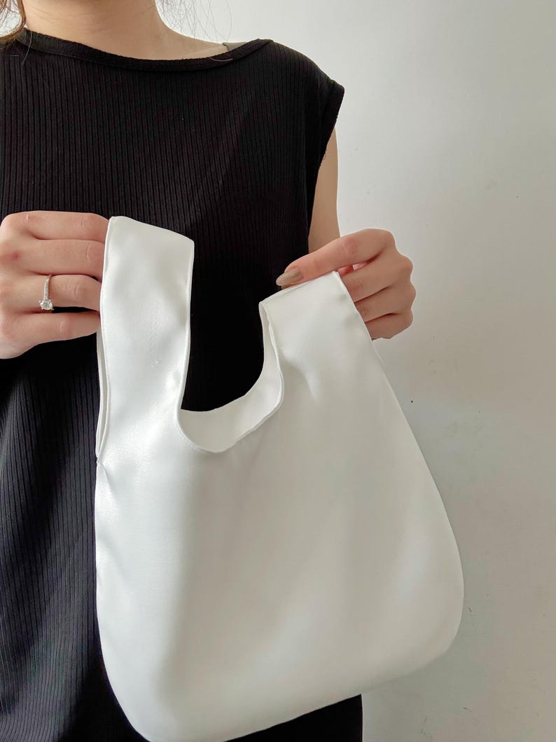 May include: A white tote bag with a rounded shape and two handles. The bag is held up by a person. The bag is made of a smooth, solid white material.