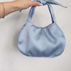 May include: A light blue satin handbag with a smooth, lustrous finish. The bag features a rounded, crescent-shaped body and a single integrated handle that is tied into a decorative knot at the top, creating a soft, gathered appearance.