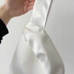 May include: A white satin knot bag featuring a smooth, lustrous finish and a single handle design. The fabric is folded into a knotted structure, and the text 10.1.26 is embroidered in white thread on the handle of the bag.