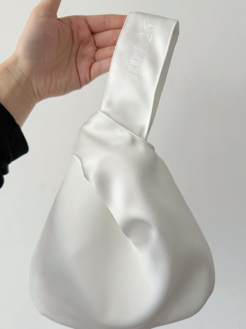 May include: A white satin knot bag featuring a smooth, lustrous finish and a single handle design. The fabric is folded into a knotted structure, and the text 10.1.26 is embroidered in white thread on the handle of the bag.