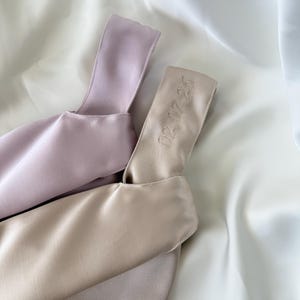 May include: Two satin bags in soft pastel colors, one lavender and one beige, rest on a white satin surface. The beige bag has the embroidered date "02.07.26" on its handle. The bags have a minimalist design.