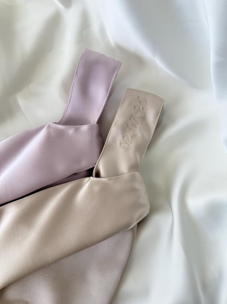 May include: Two satin bags in soft pastel colors, one lavender and one beige, rest on a white satin surface. The beige bag has the embroidered date "02.07.26" on its handle. The bags have a minimalist design.