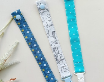 May include: Three fabric pacifier clips featuring distinct patterns: a dark blue strap with yellow and white stars, a white strap with black line-art illustrations of cartoon characters, and a bright turquoise strap with a white cross-stitch pattern.