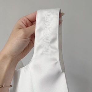 May include: A white satin fabric strap featuring the embroidered date 06.06.2026 in a matching white thread. The smooth, lustrous material is shown in a vertical orientation, highlighting the clean, tonal stitching of the numerical text.