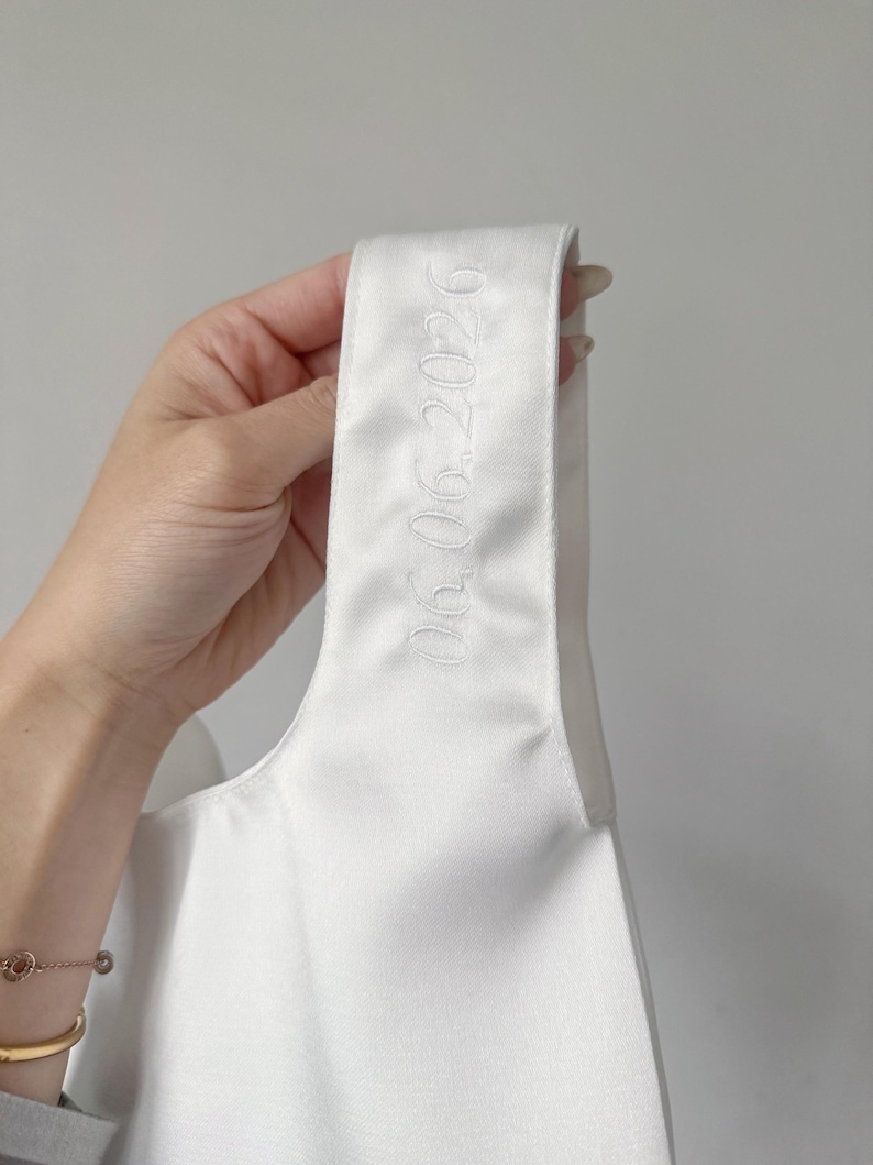 May include: A white satin fabric strap featuring the embroidered date 06.06.2026 in a matching white thread. The smooth, lustrous material is shown in a vertical orientation, highlighting the clean, tonal stitching of the numerical text.