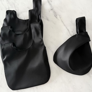 May include: Four black satin tote bags of varying sizes are displayed on a marble surface. The bags have a simple, minimalist design with a knotted handle. The bags are empty and appear to be made of a smooth, shiny fabric.