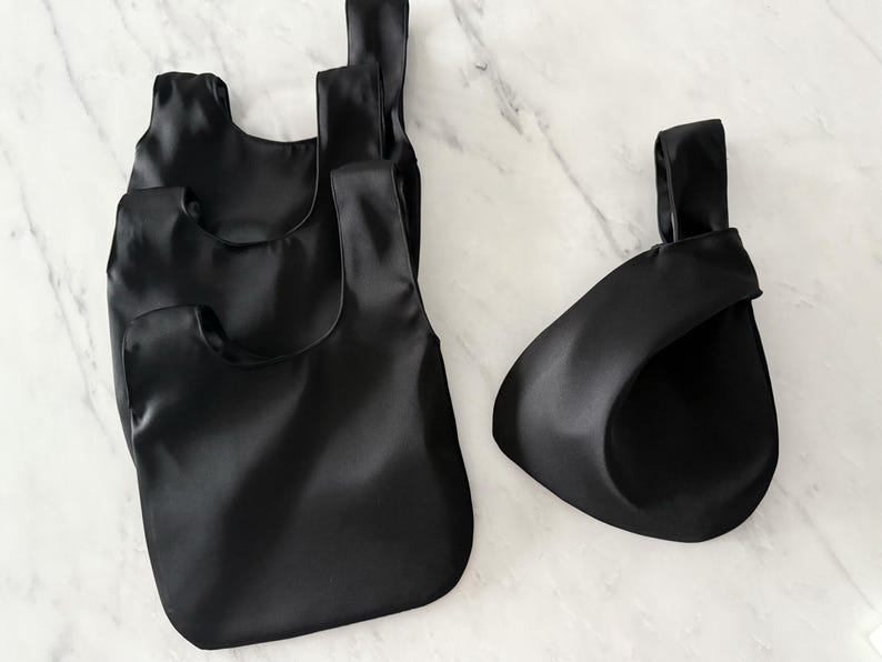 May include: Four black satin tote bags of varying sizes are displayed on a marble surface. The bags have a simple, minimalist design with a knotted handle. The bags are empty and appear to be made of a smooth, shiny fabric.