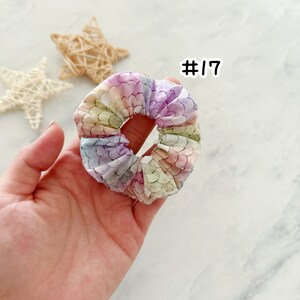 K&ouml;nnte beinhalten: Ein pastellfarbener Regenbogen-Scrunchie mit einem Meerjungfrauen-Schuppenmuster. Der Scrunchie wird in einer Hand gehalten und die Nummer #17 ist dar&uuml;ber geschrieben.
