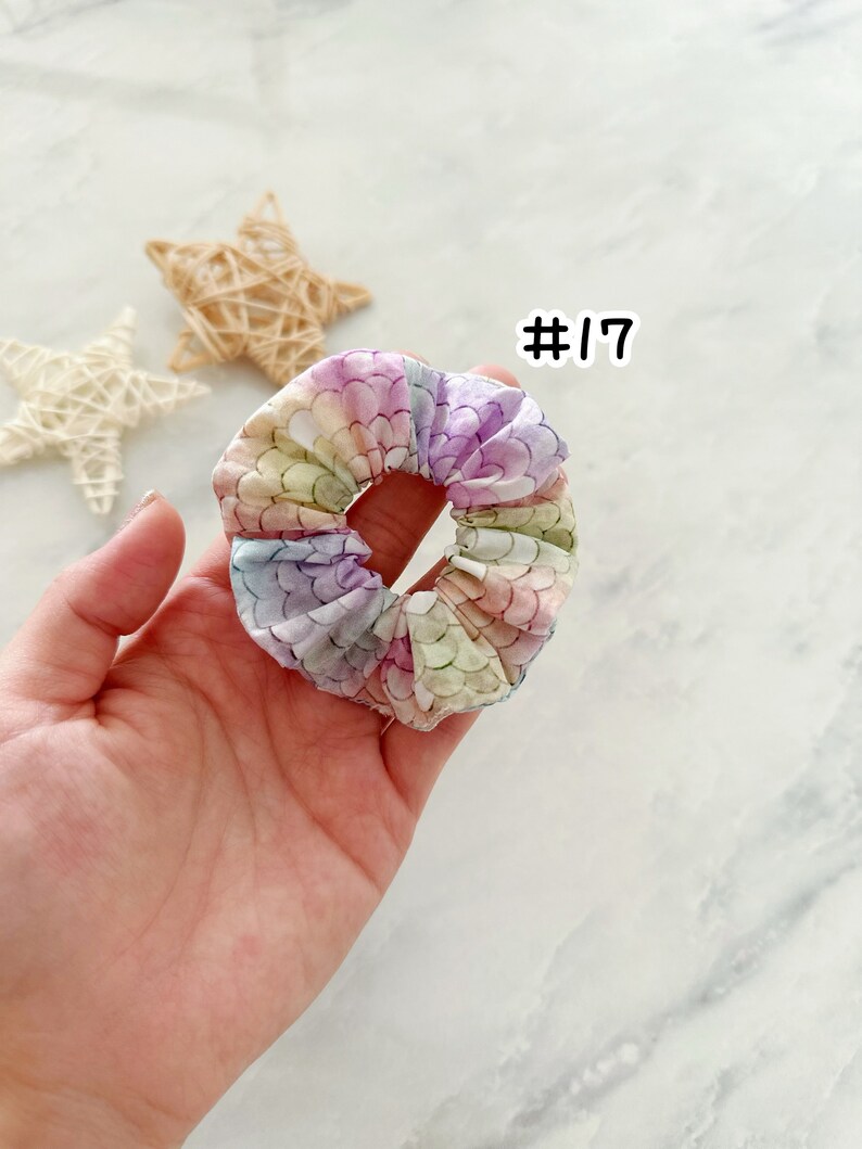 K&ouml;nnte beinhalten: Ein pastellfarbener Regenbogen-Scrunchie mit einem Meerjungfrauen-Schuppenmuster. Der Scrunchie wird in einer Hand gehalten und die Nummer #17 ist dar&uuml;ber geschrieben.
