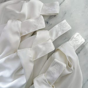 May include: Several white satin bags with embroidered initials on the handle. The bags are arranged on a marble surface. The initials are in a delicate script, and the bags appear to be for a special occasion.