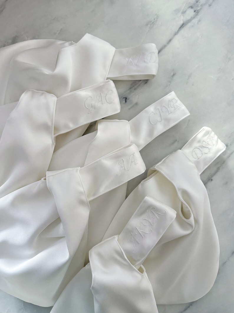 May include: Several white satin bags with embroidered initials on the handle. The bags are arranged on a marble surface. The initials are in a delicate script, and the bags appear to be for a special occasion.
