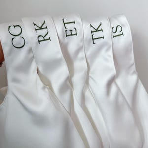 May include: Five white satin bags with handles, each featuring dark green embroidered initials. The initials displayed on the bags are CG, RK, ET, TK, and IS, arranged in a row against a plain, light-colored background.