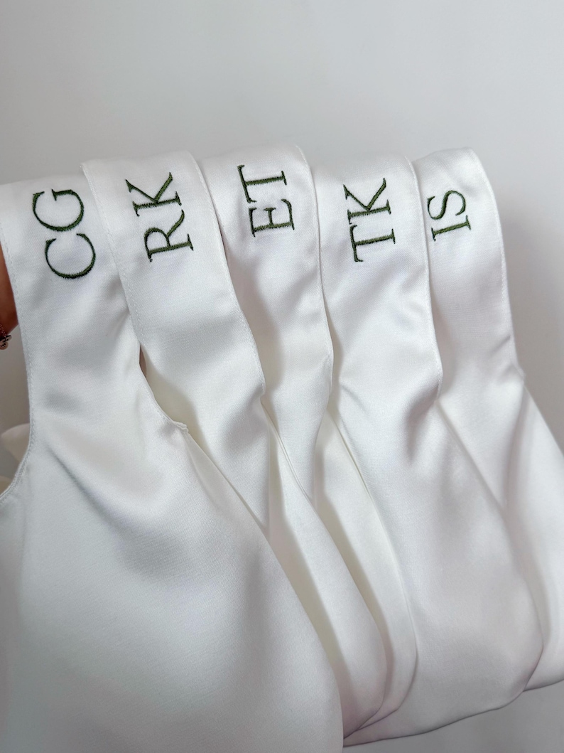 May include: Five white satin bags with handles, each featuring dark green embroidered initials. The initials displayed on the bags are CG, RK, ET, TK, and IS, arranged in a row against a plain, light-colored background.
