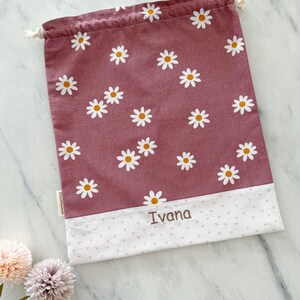 May include: A pink drawstring bag with a white daisy pattern and a white bottom with a pink heart pattern. The bag has the name "Ivana" embroidered on the bottom.