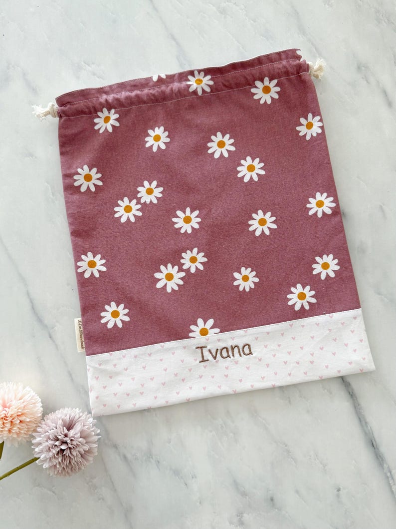 May include: A pink drawstring bag with a white daisy pattern and a white bottom with a pink heart pattern. The bag has the name "Ivana" embroidered on the bottom.