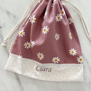 May include: A pink drawstring bag with a white daisy pattern and a white bottom with a pink heart pattern. The bag has the name "Clara" embroidered on the bottom.