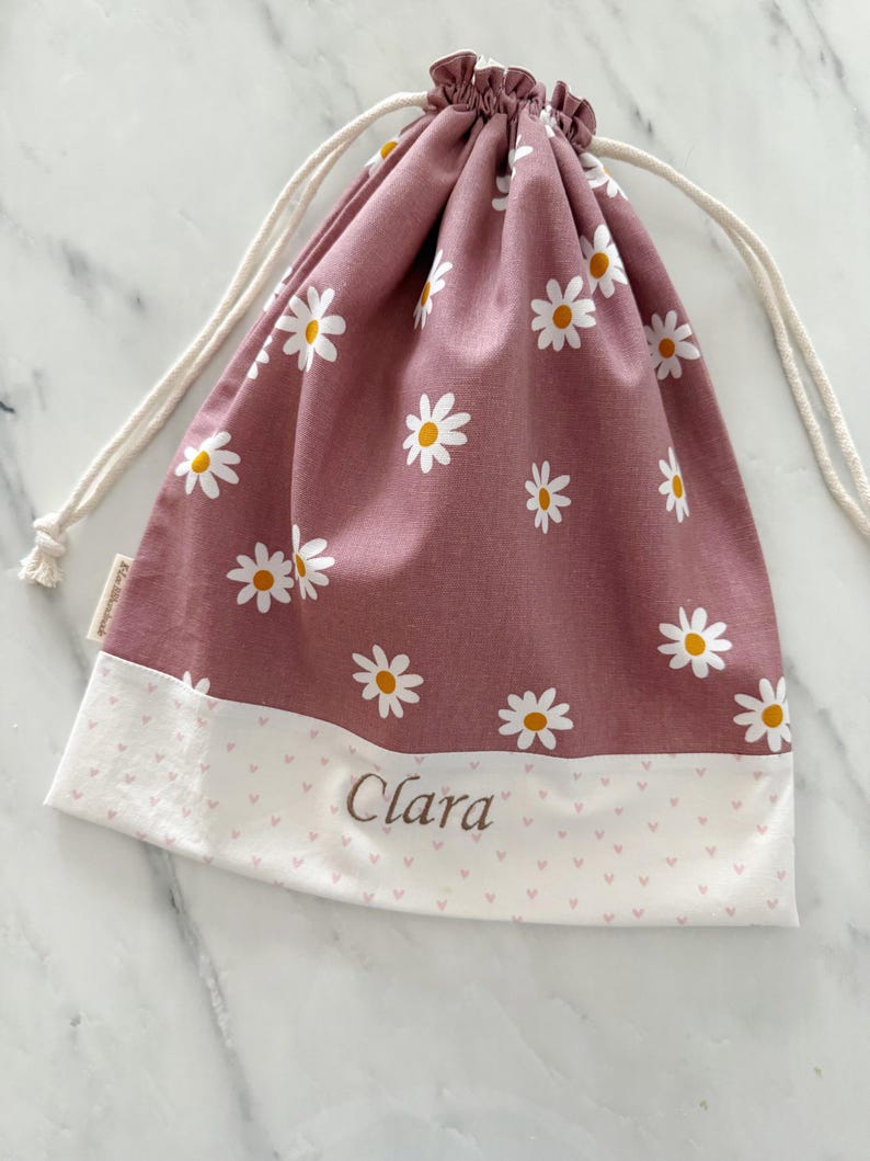 May include: A pink drawstring bag with a white daisy pattern and a white bottom with a pink heart pattern. The bag has the name "Clara" embroidered on the bottom.