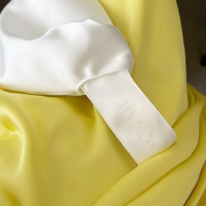 May include: Close-up of a white satin garment with a yellow satin background. A white satin tag is attached to the garment, with the date "10.1.26" embroidered on it. The fabric has a smooth, shiny texture.