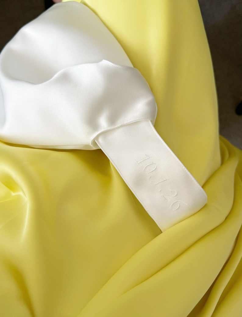 May include: Close-up of a white satin garment with a yellow satin background. A white satin tag is attached to the garment, with the date "10.1.26" embroidered on it. The fabric has a smooth, shiny texture.