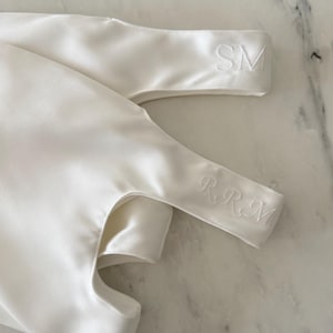 May include: A smooth white satin fabric item featuring two distinct embroidered sections. One section displays the letters SM in a clean font, while the other section features the letters RRM in an elegant, cursive-style script embroidery.