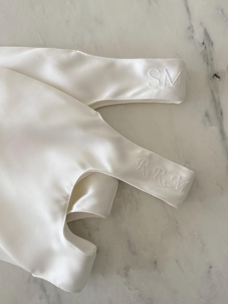 May include: A smooth white satin fabric item featuring two distinct embroidered sections. One section displays the letters SM in a clean font, while the other section features the letters RRM in an elegant, cursive-style script embroidery.