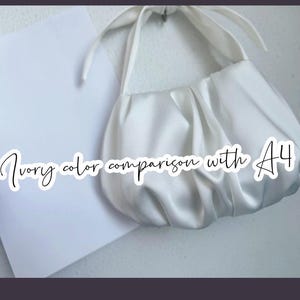 May include: An ivory-colored, gathered satin handbag with a knotted handle, positioned next to a standard A4 sheet of white paper. The text "Ivory color comparison with A4" is overlaid on the image in a white script font.