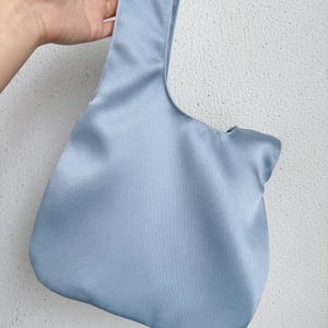 May include: A light blue satin tote bag with a single handle. The bag has a rounded bottom and a smooth, shiny texture. The handle is wide and integrated into the bag's design. The bag is held against a white textured wall.