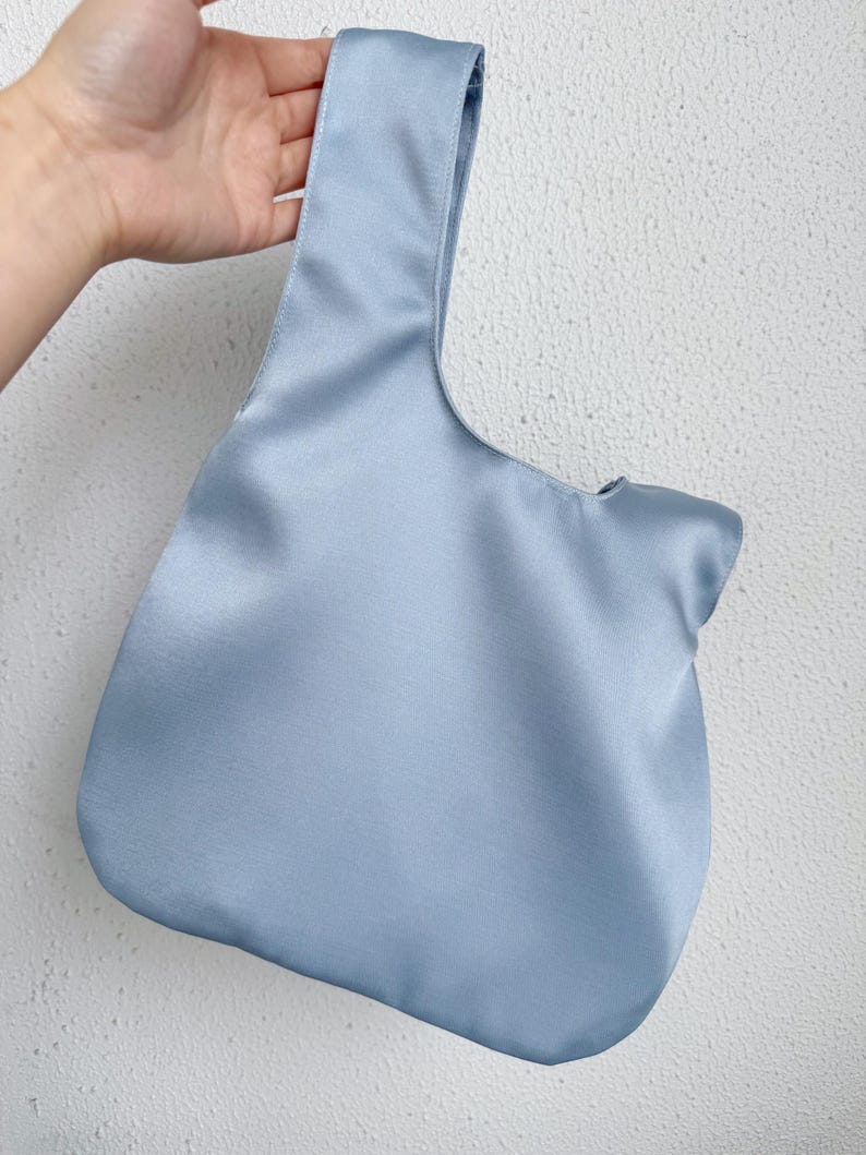 May include: A light blue satin tote bag with a single handle. The bag has a rounded bottom and a smooth, shiny texture. The handle is wide and integrated into the bag's design. The bag is held against a white textured wall.
